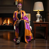 Woman sitting in a chair wearing a colorful Bird of Paradise Pink Purple Silk Headscarf and Kimono in a cozy room with a fireplace.