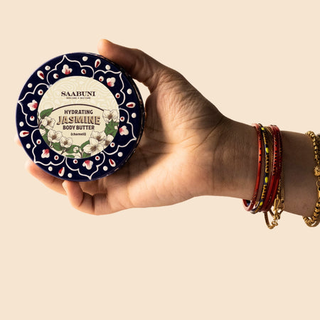 Hand holding a container of Saahuni Hydrating Jasmine Body Butter against a beige background