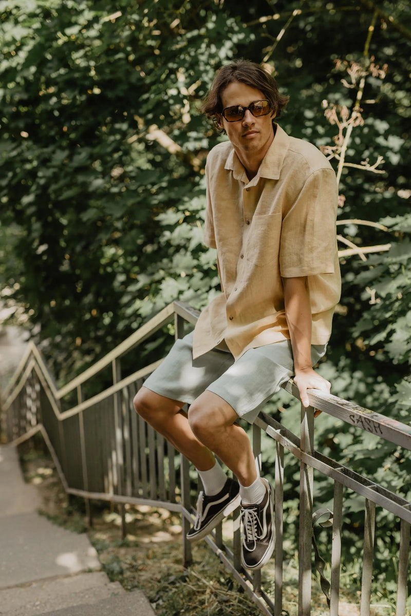 Man Sitting On Railing Wearing A Shirt And Sage Green Linen Shorts