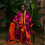 Man wearing a vibrant, multicolored Bird of Paradise Pink Purple Silk Kimono and orange pants sitting on a chair with plants in the background.