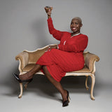 Woman in a Boudicca Tomato Red Ruched Midi Dress sitting on an ornate chair against a gray background