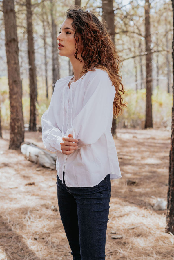 Mesa blouse in cloud white color for women by Paneros Clothing. Handmade from sustainable deadstock cotton poplin fabric, button-up with full sleeves, tie neck, and slight shirttail. Side view.